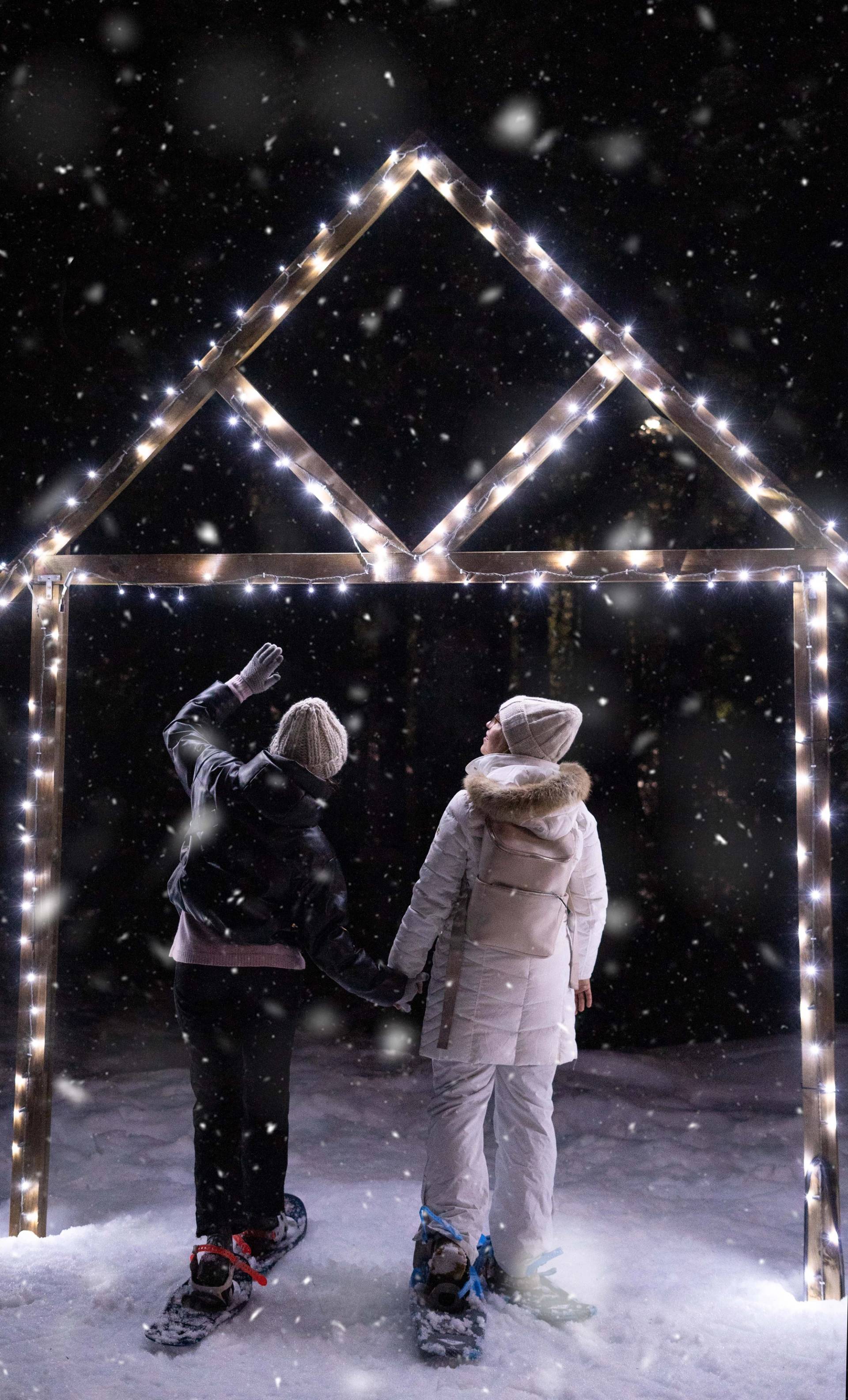 Two people snowshoe while looking up at lights at night.