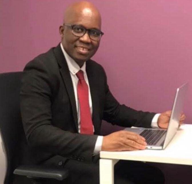 Mamanding “Max” Daffeh at his desk with a laptop, in a suit and tie, smiling at the camera.