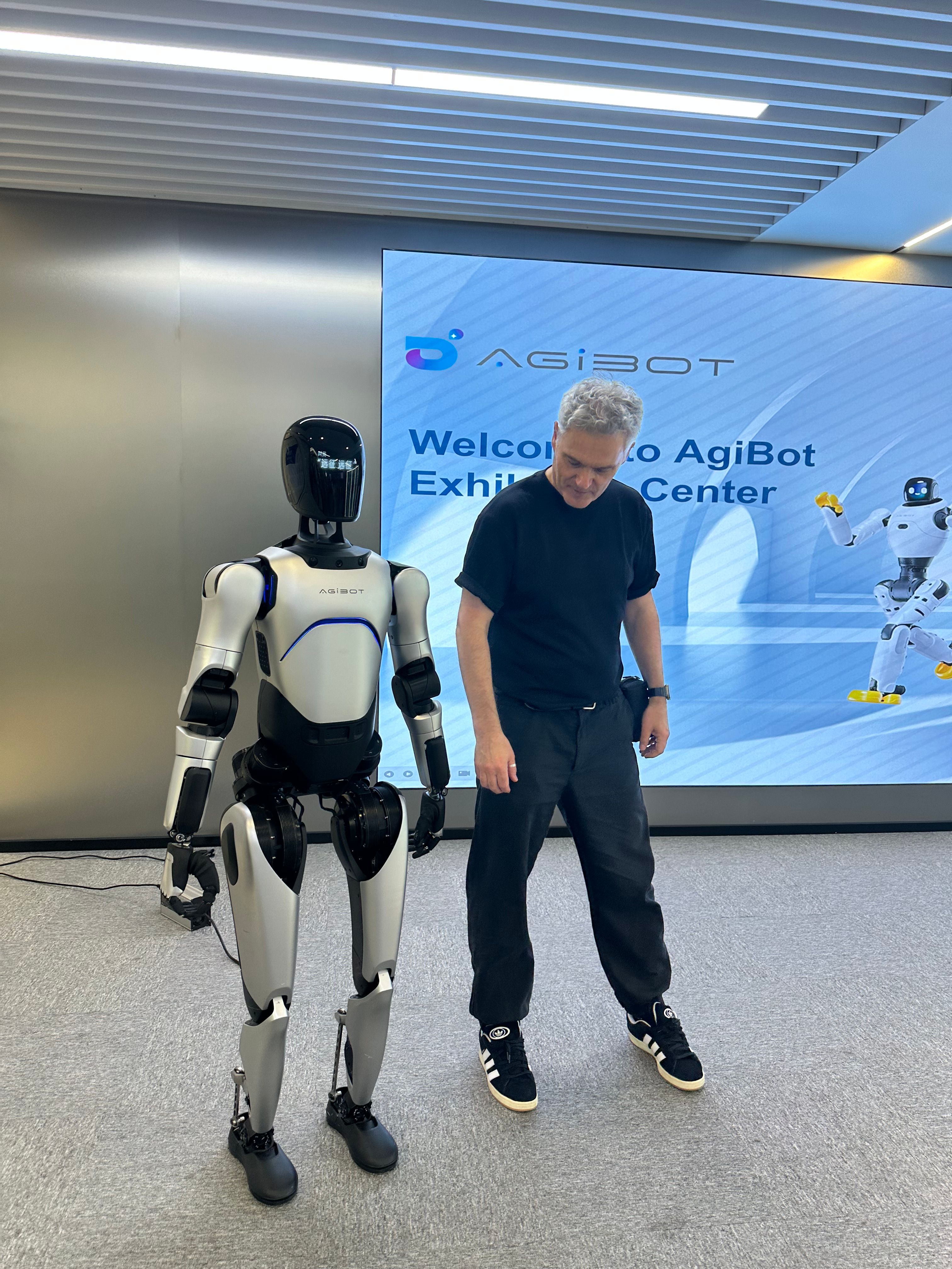 Ein humanoider Roboter steht neben Erik Rippmann in einem Ausstellungsraum eines Robotik-Startups in China; im Hintergrund ist ein Display mit dem Schriftzug ‚Welcome to AgiBot Exhibition Center