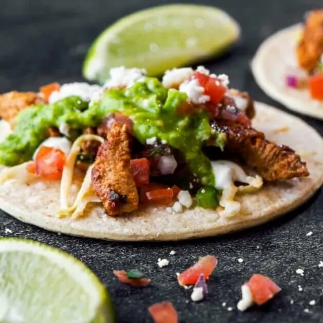 Tacos topped with salsa and guacamole beside lime wedges.