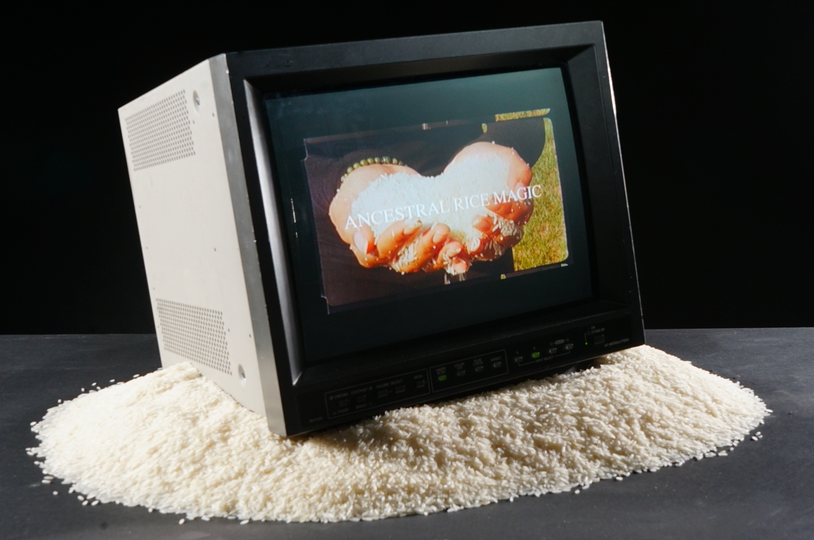 CRT monitor on a small mountain of rice.  Screen showing title of video with hands holding rice 