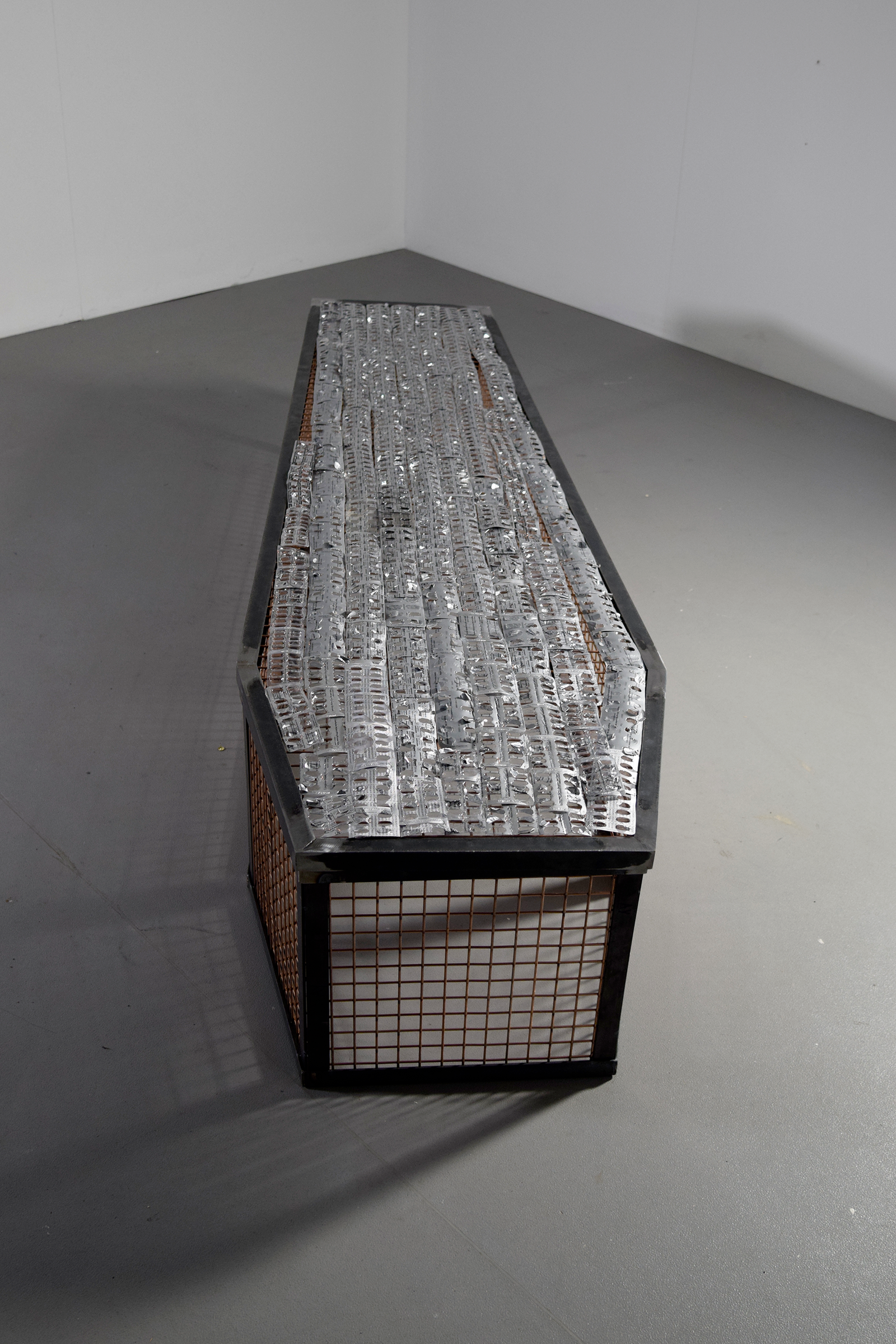 Photo of a life-sized coffin-shaped cage covered in empty used blister packets.