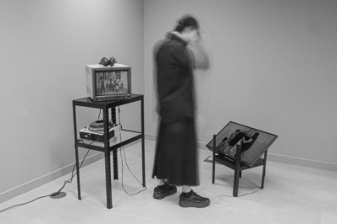 Black and white photo of a room with two television screens on furniture, blurred person watching
