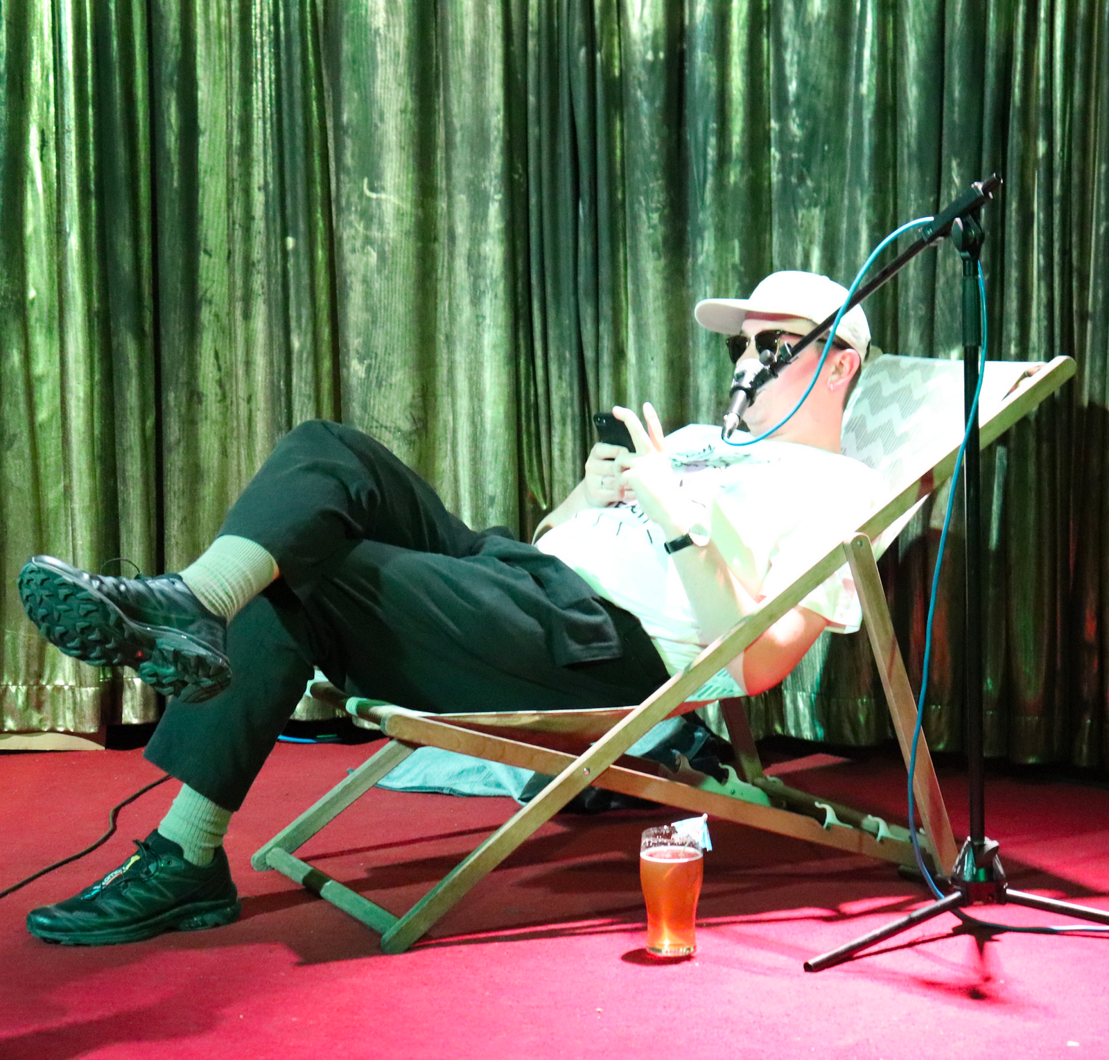 Artist Ben Sargent sitting in a deckchair on a stage reading a piece of writing from his phone.