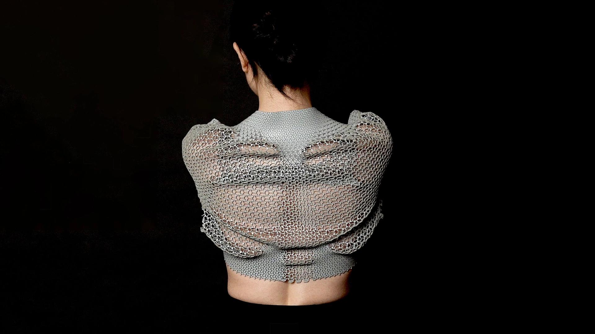 Photograph of a female figure, back to camera, with grey geometric cutout mesh top, black background
