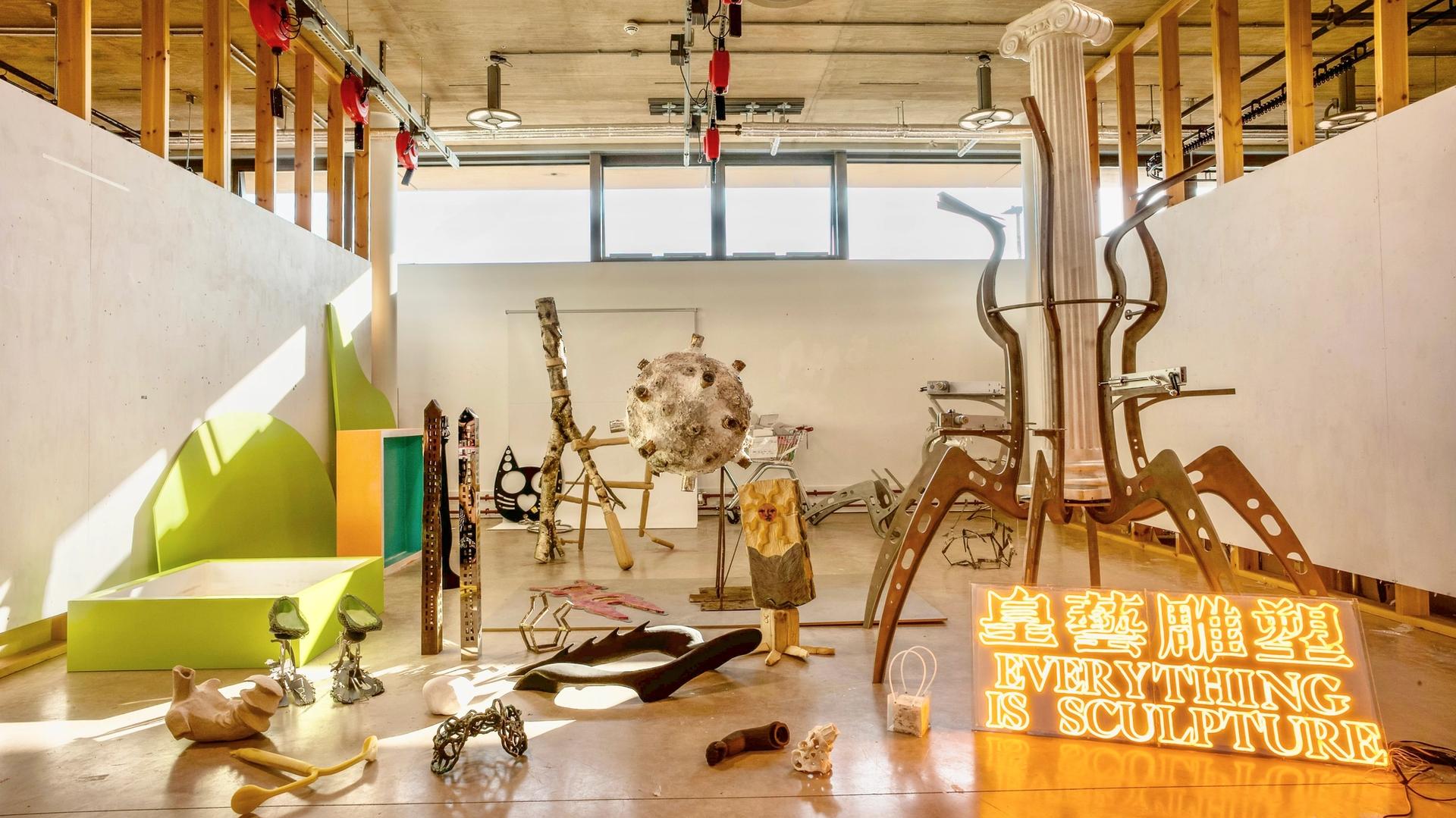 A photograph of a corner of a large space filled with sculptural works and various works on the floor and attached to a wall.