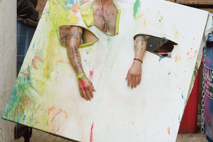 A person standing with a canvas that has been placed over their head and over their arms. At their feet are bottles of paint and the ground is covered in green paint.