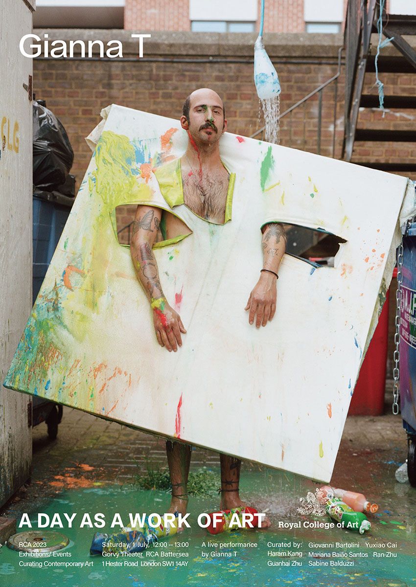 A person standing with a canvas that has been placed over their head and over their arms. At their feet are bottles of paint and the ground is covered in green paint.