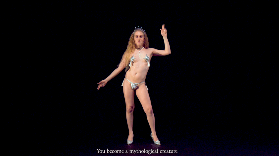 A white trans person with long curly hair is wearing a tiara, pasties and a thong while dancing in front of a black background.