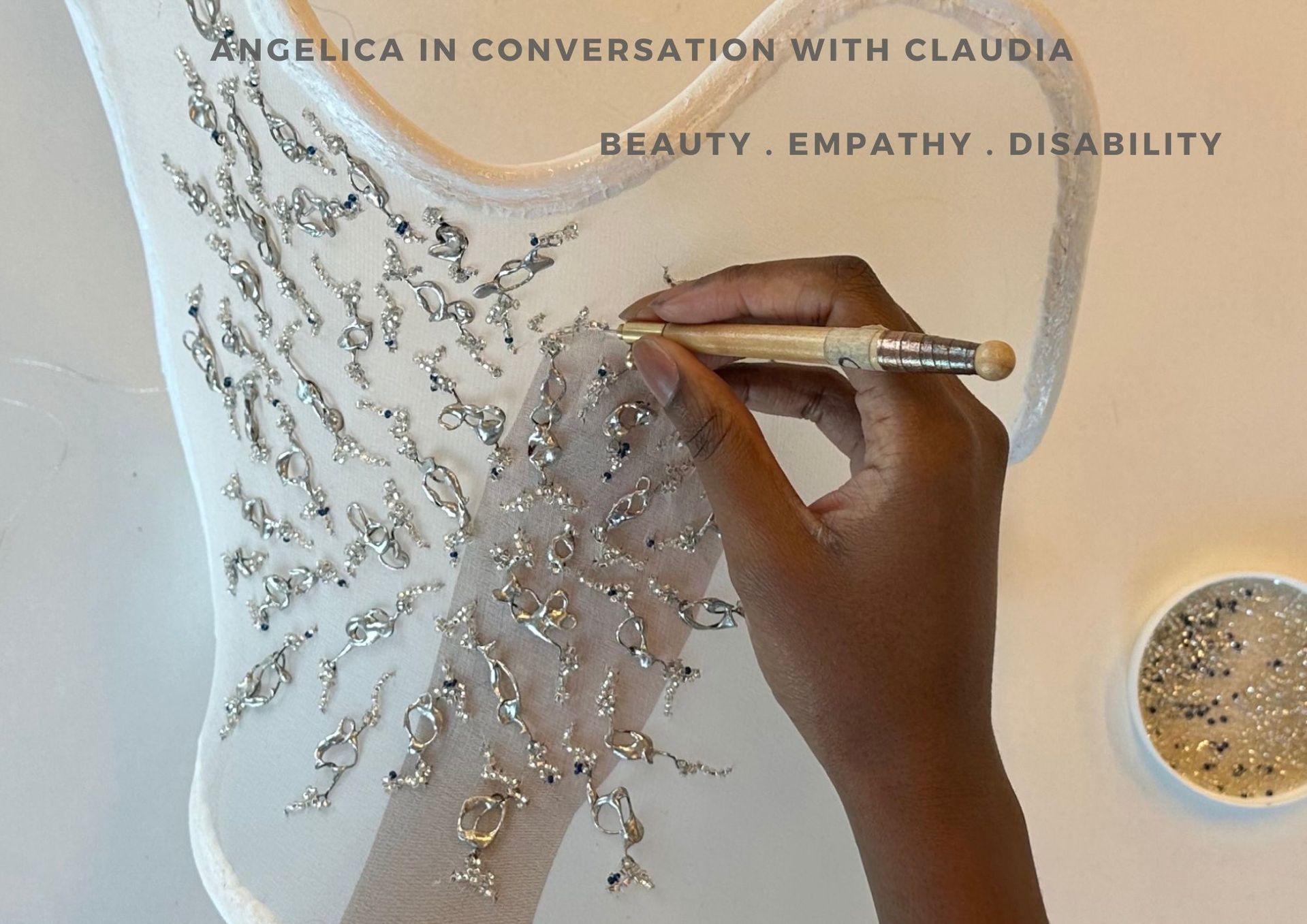 Image of a pair of hands embroidering beads onto what looks like a corset made out of sheer fabric. The sheer fabric is pale ivory and the beads are silver and irregularly shaped. 