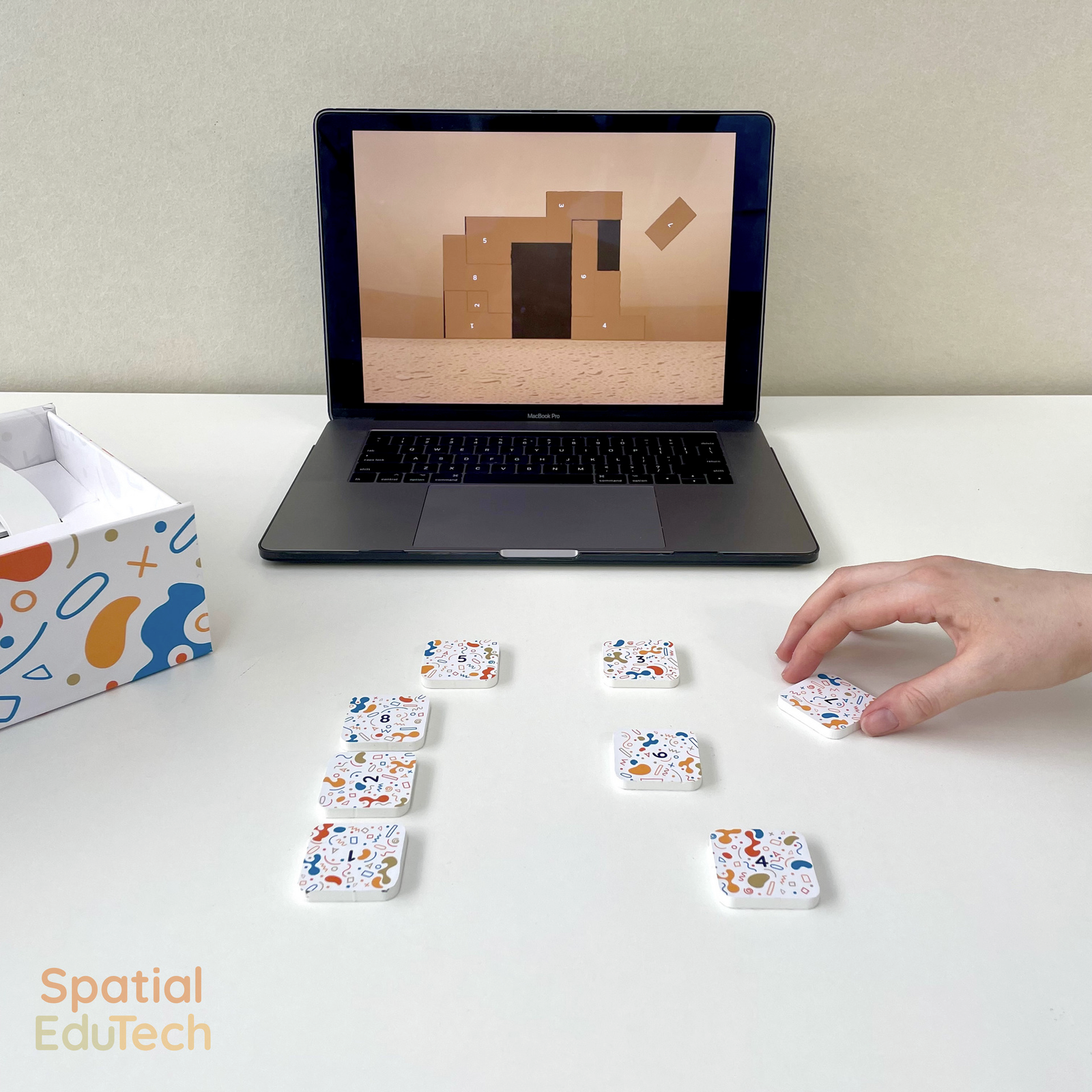 A tiling puzzle on a laptop with physical game pieces in front, with a hand positioning a piece to solve the digital puzzle.