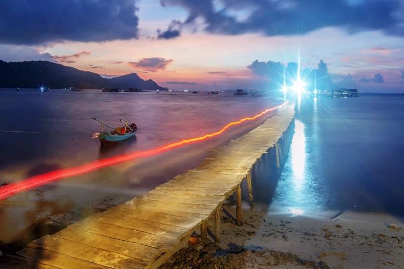 Evening light trails along a pier with boats and a darkening coastline