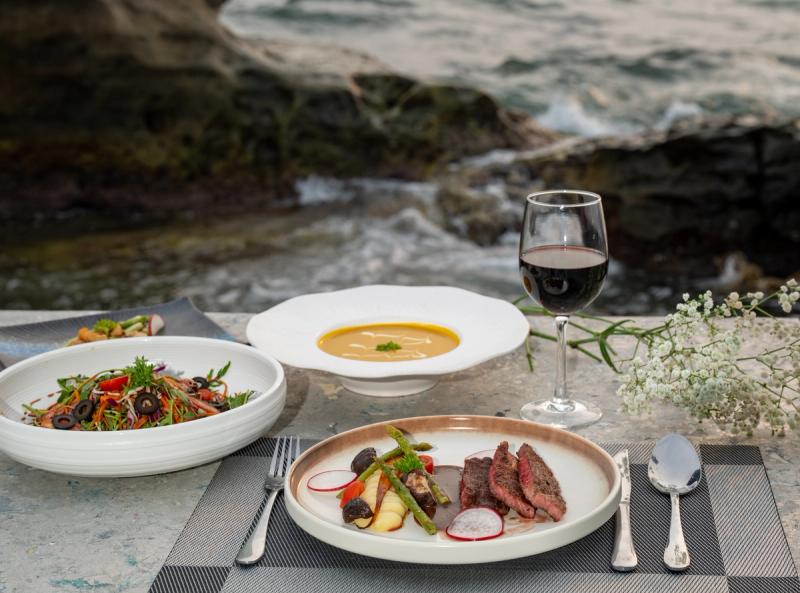 Plated dishes and a wine glass on a table beside coastal rocks