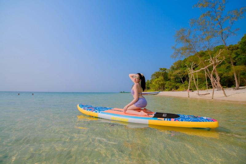 A person kneeling on a paddleboard beside a tree-lined beach