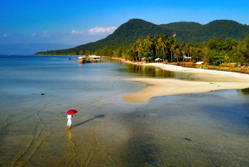 A person carrying a red umbrella beside the Rach Vem shore and wooded hills