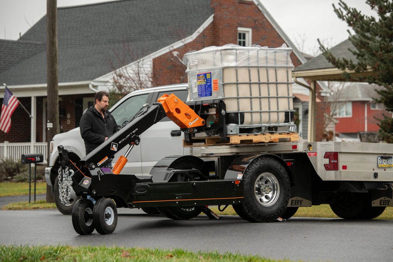 An Equipter Tow-A-Lift lifts a palletized tote off the back of a flatbed truck on a residential street, with the operator standing alongside.