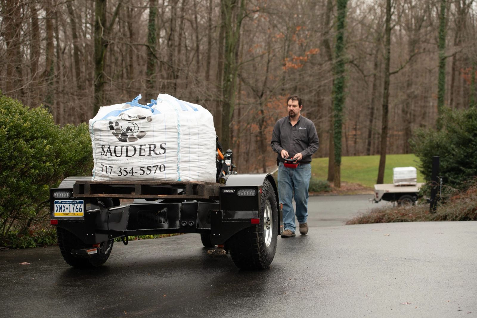 An Equipter Tow-A-Lift carries a palletized bulk bag up a wet residential driveway while the operator walks alongside holding the handheld remote.
