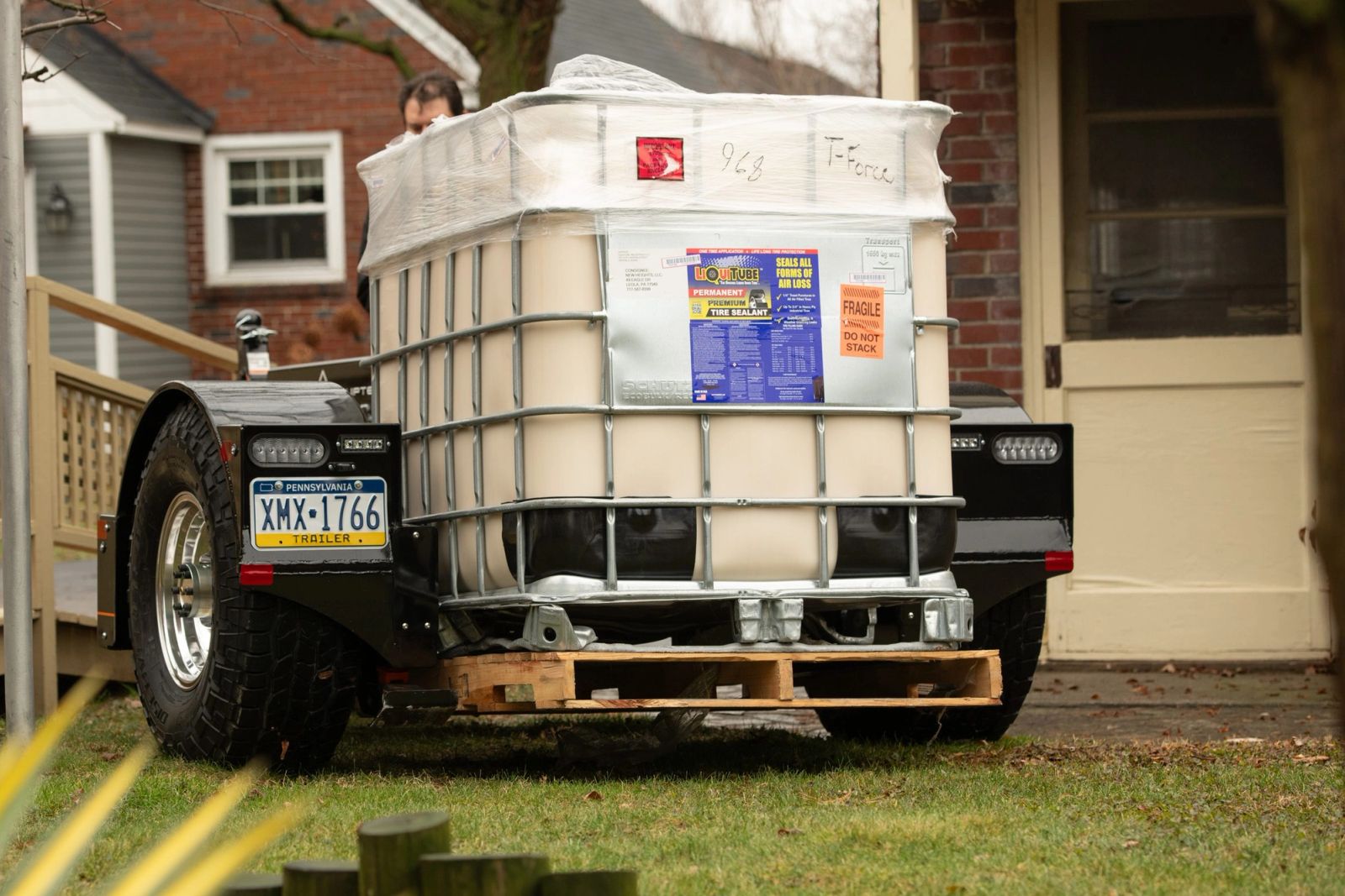An Equipter Tow-A-Lift sets a palletized IBC tote down beside a brick house in a residential back yard, working between the building and a wooden ramp.