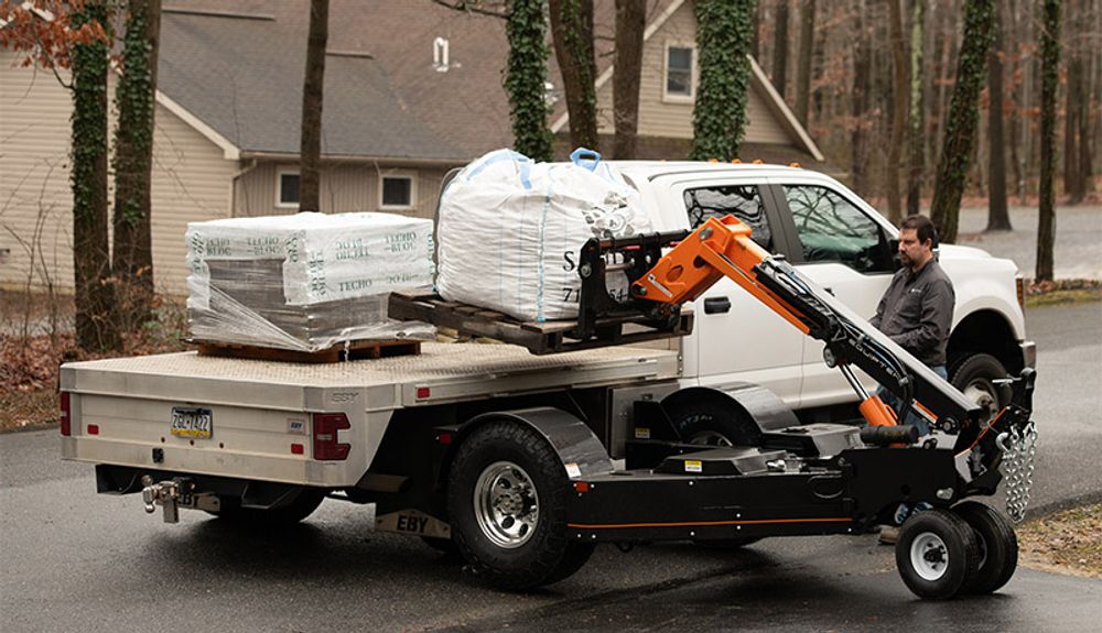 An Equipter Tow-A-Lift reaches onto the bed of a flatbed truck to pick up a pallet of pavers and a bulk bag, with the operator standing at the side of the truck.