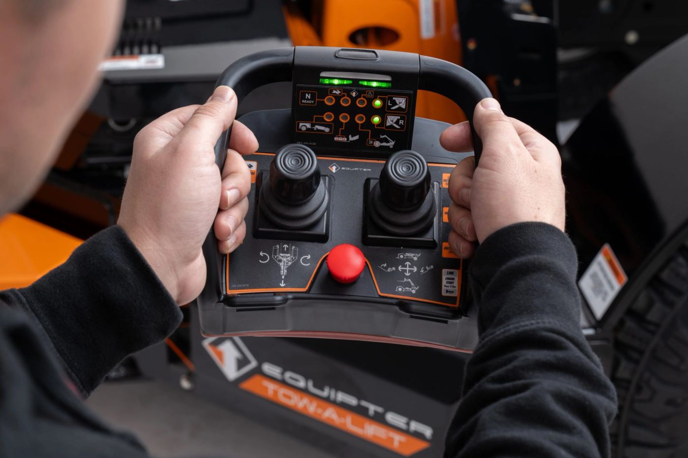 An operator's hands on the Equipter Tow-A-Lift wireless remote, with dual joysticks and a red emergency stop.
