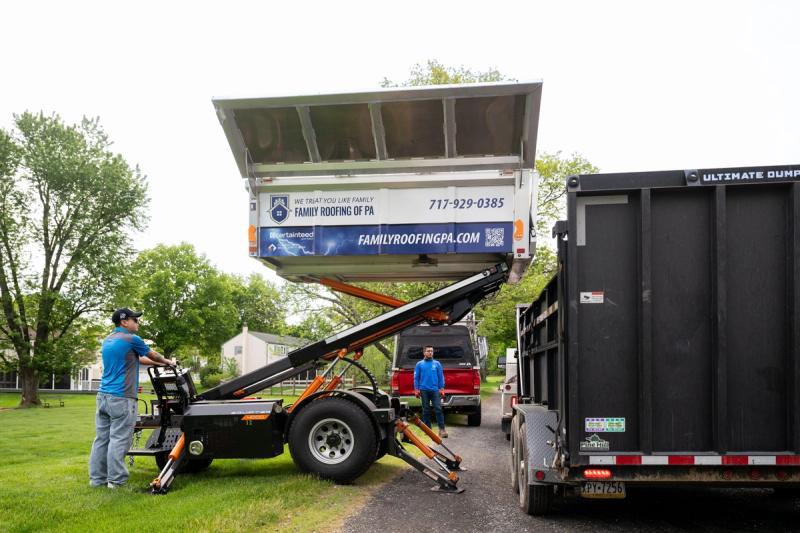 An Equipter 4000 lifting and tipping a load of roofing debris into a dump trailer at the curb.