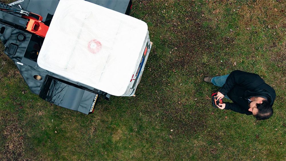 Overhead view of an Equipter Tow-A-Lift carrying a large white tote across a grass lawn while the operator walks alongside with the remote.