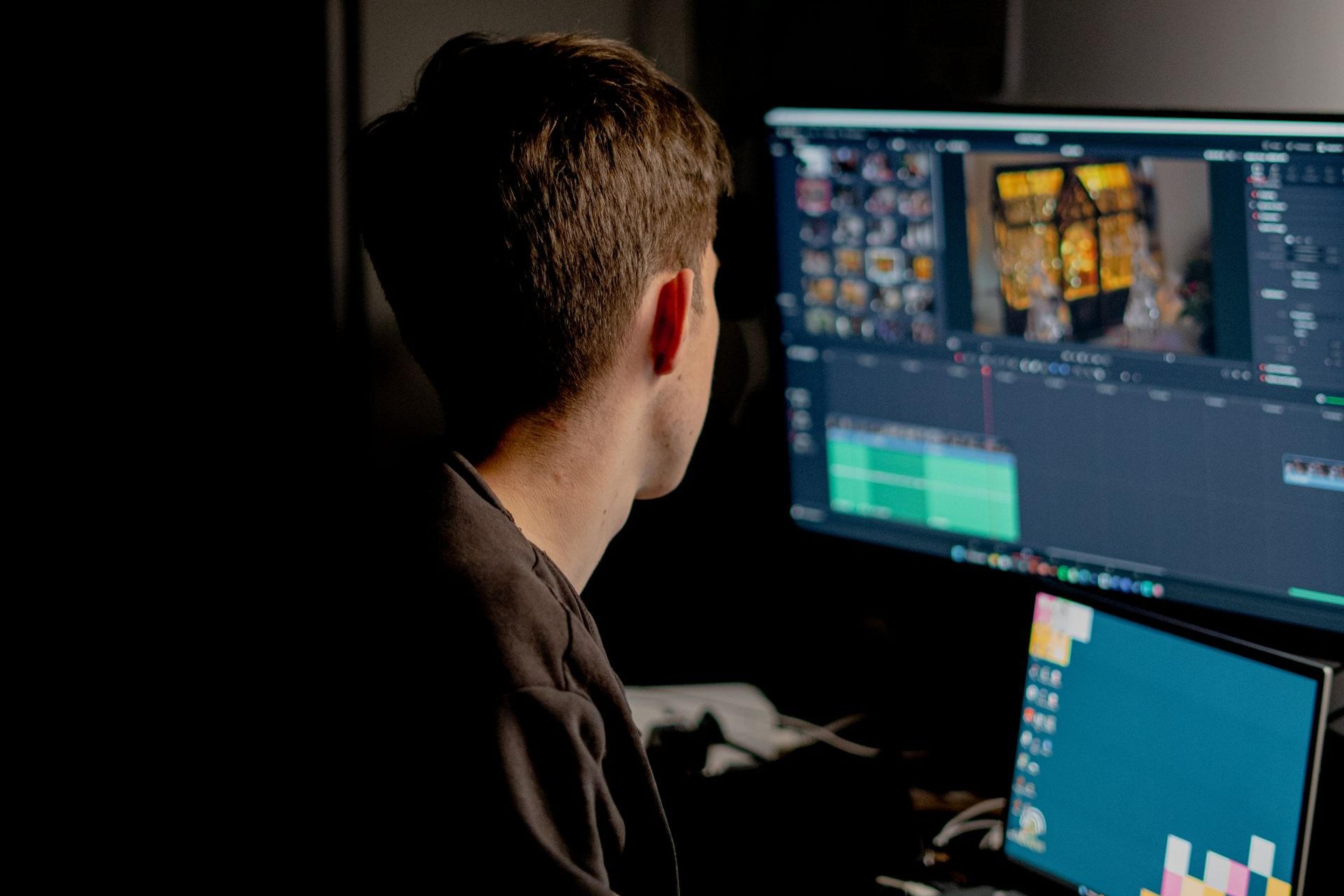 Editor at a dual-monitor workstation in a darkened suite, a video timeline open on screen