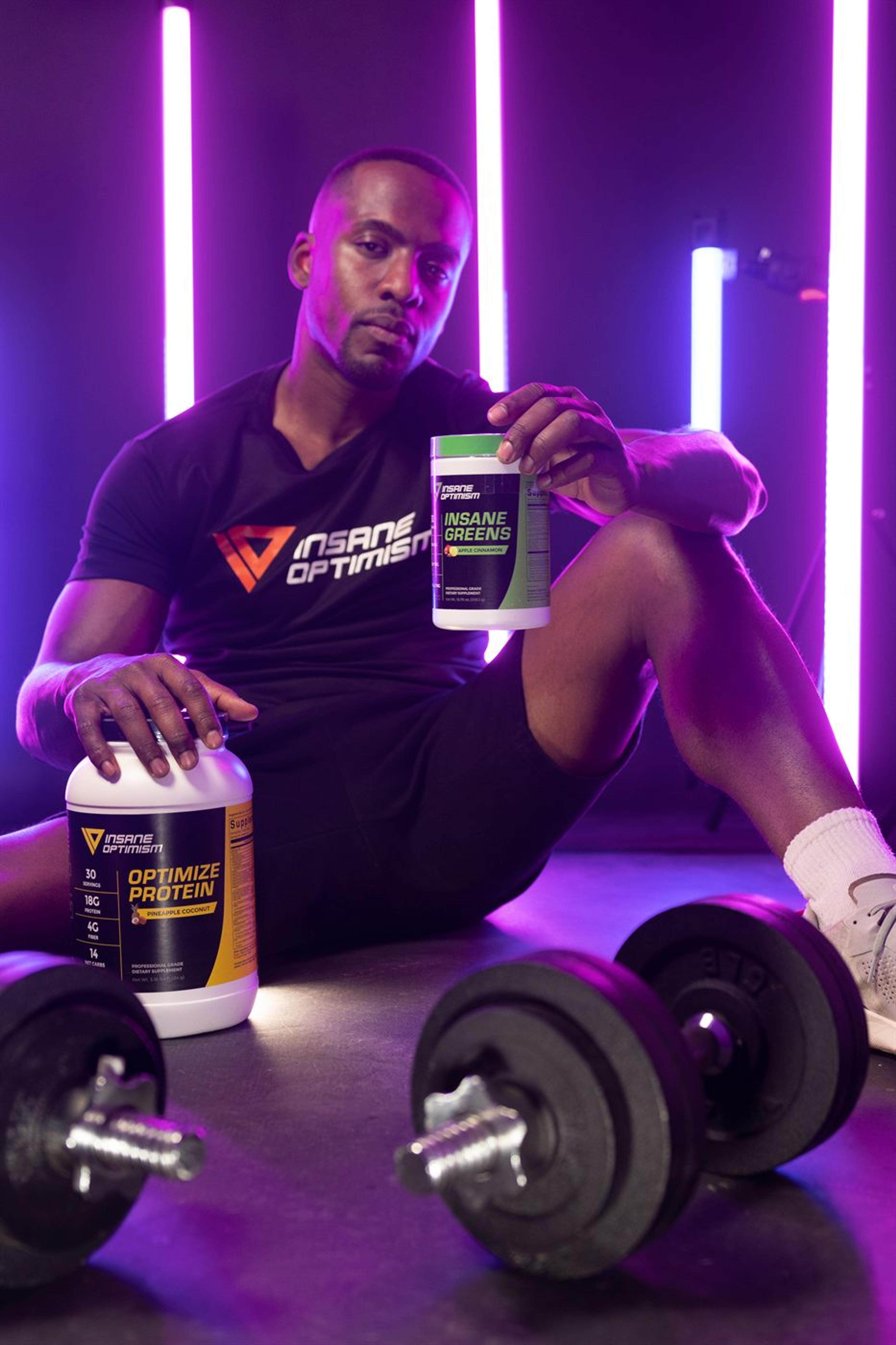 Athlete seated between magenta neon tubes holding a supplement tub beside a dumbbell rack