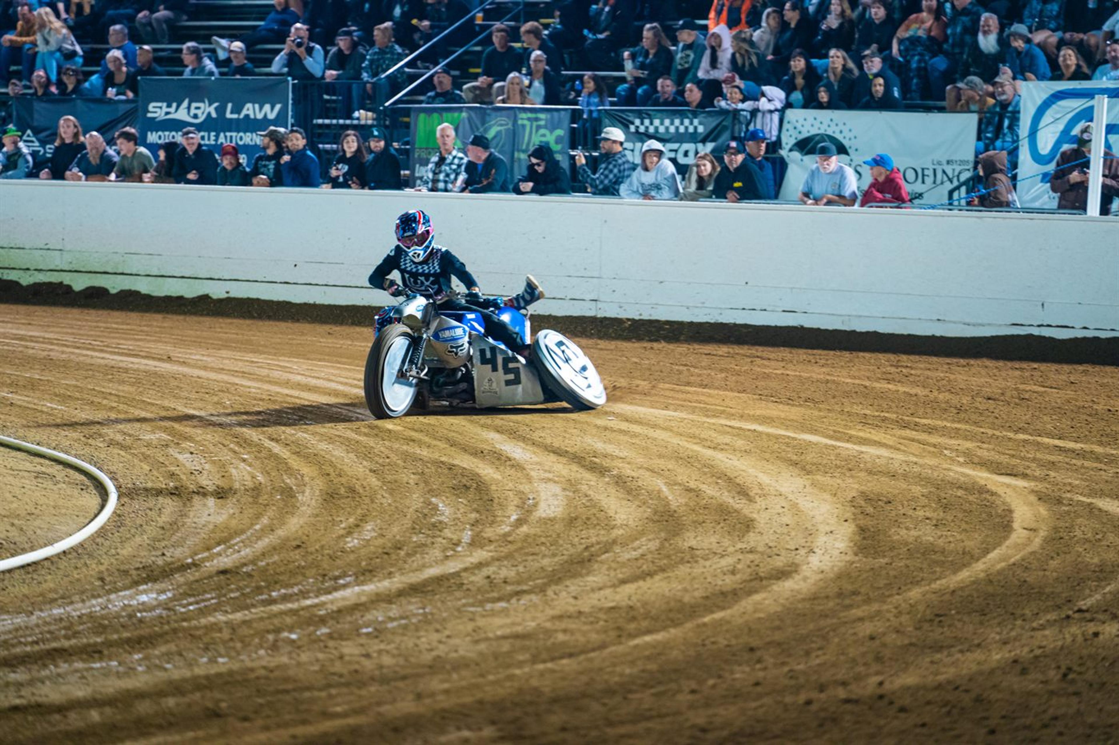 Flat track motorcycle racer leaning into a dirt corner in front of a grandstand