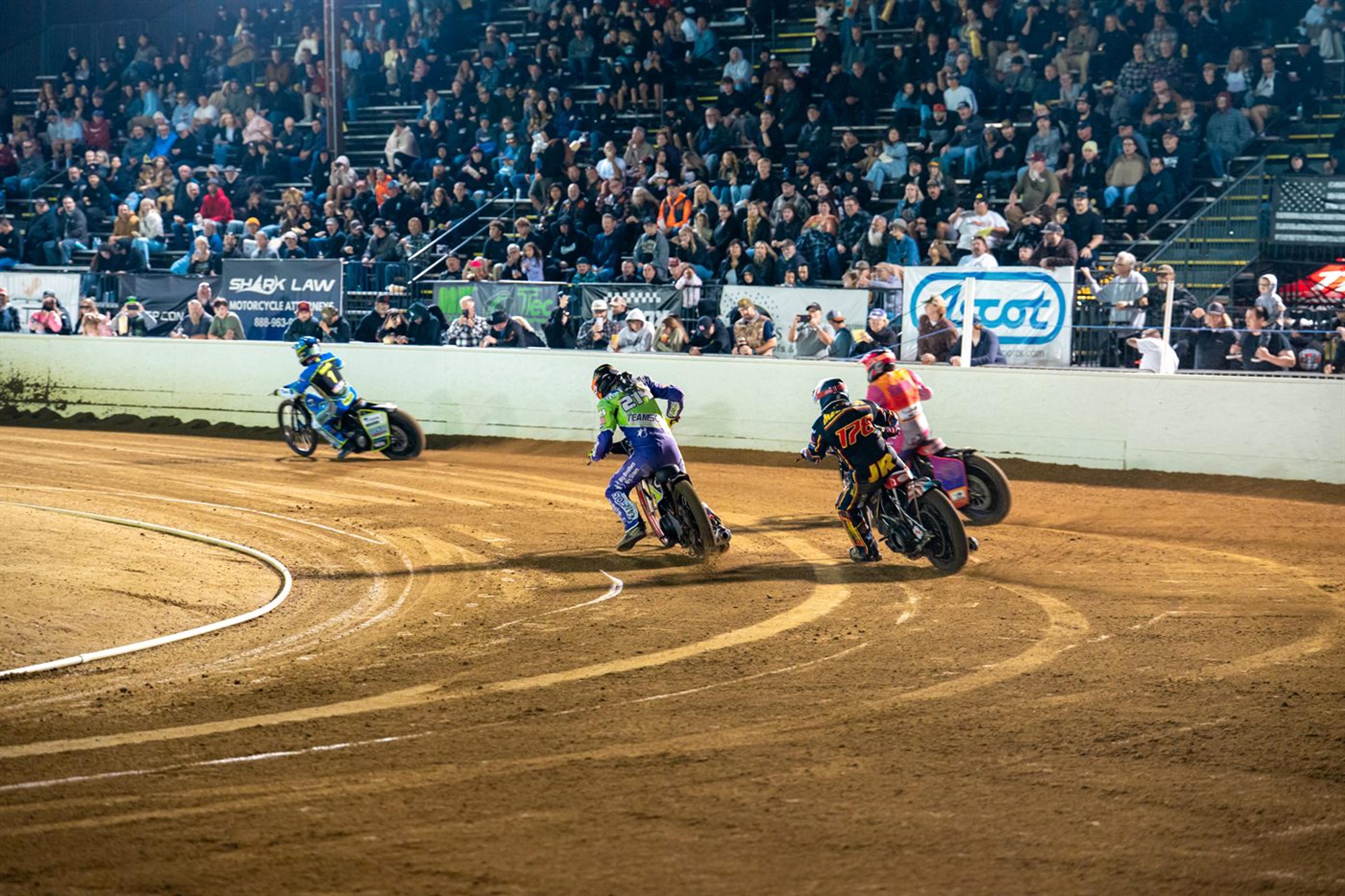 Three flat track racers leaning through a dirt corner in front of a packed grandstand