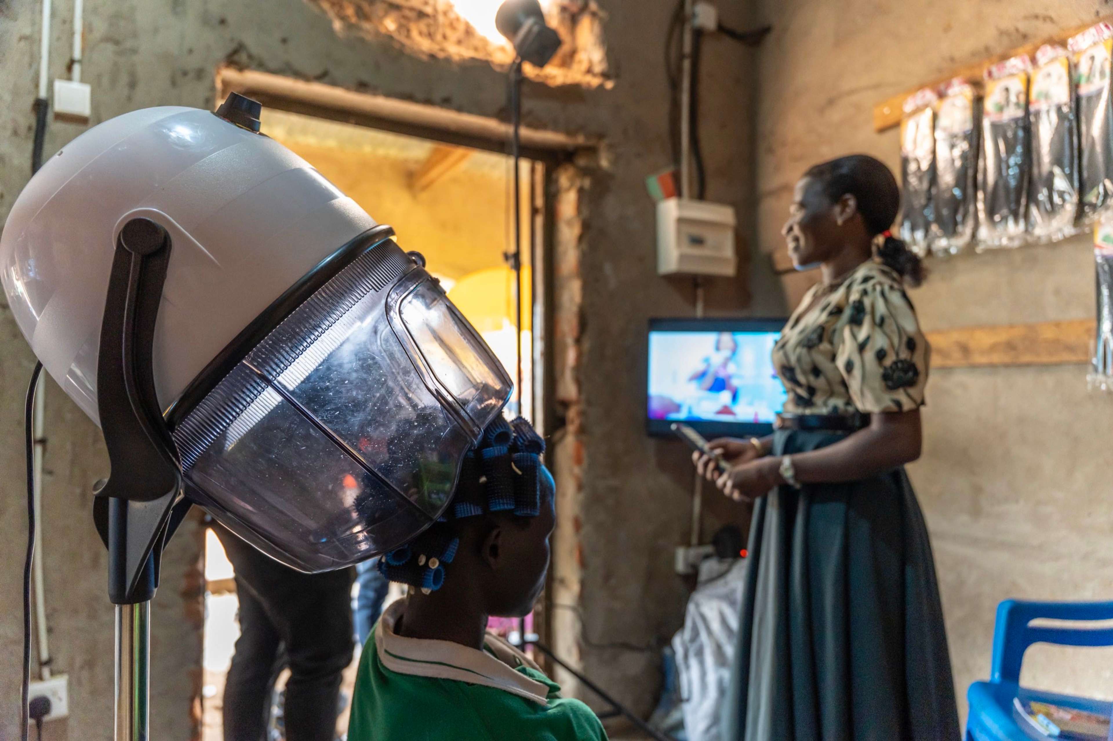 A client sits under a hair dryer in a salon, while a woman holding a phone stands nearby with hair extensions on the wall.