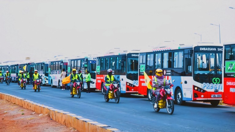 A line of motorcyclists in yellow vests on red bikes leads a procession of white, blue, and red buses on a road.