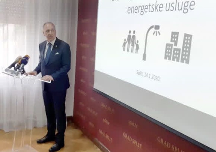 A man in a suit speaks at a podium with microphones, next to a screen showing icons of people, a street light, and buildings under the title "energetske usluge."