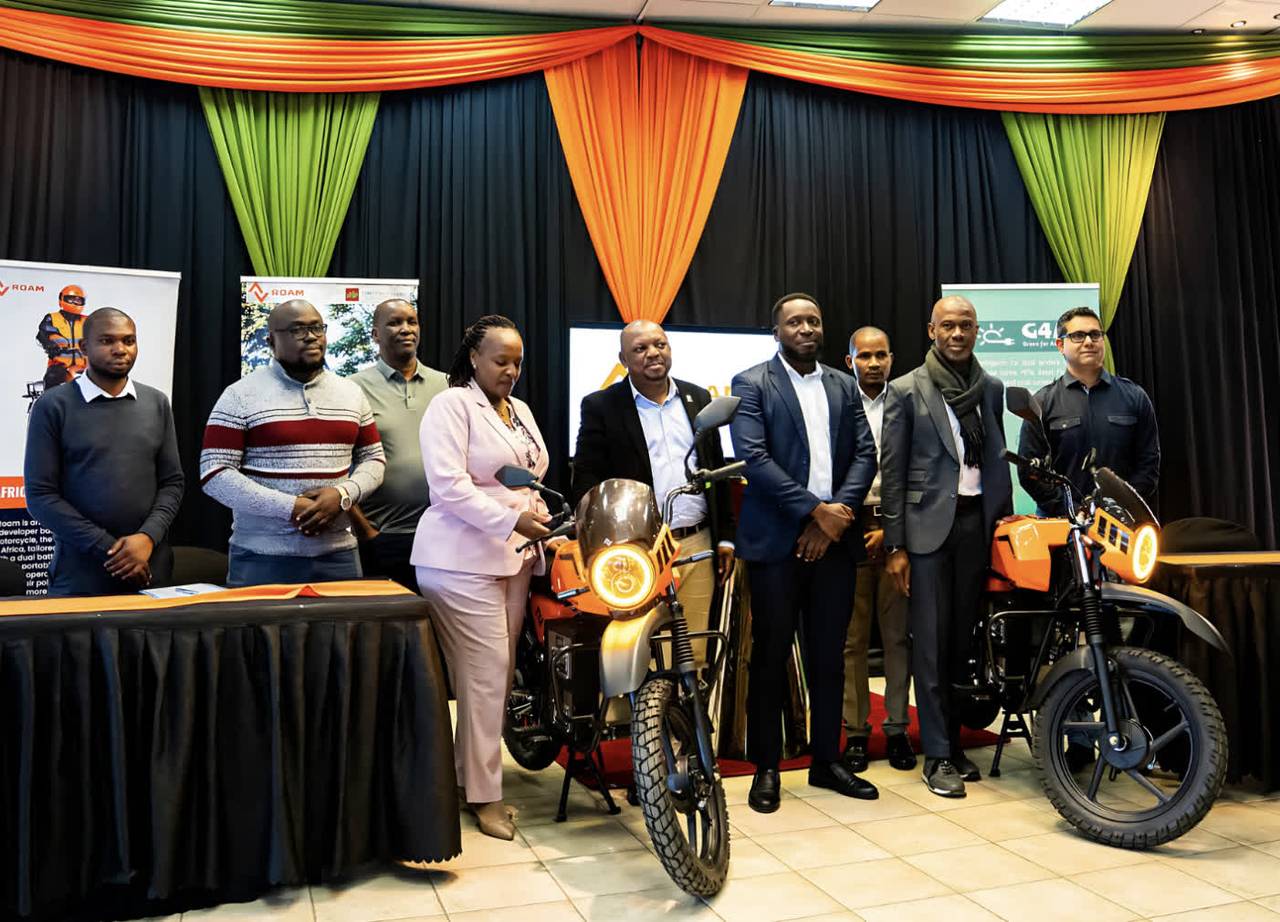 A group of professionals stands with two orange motorcycles at an event.