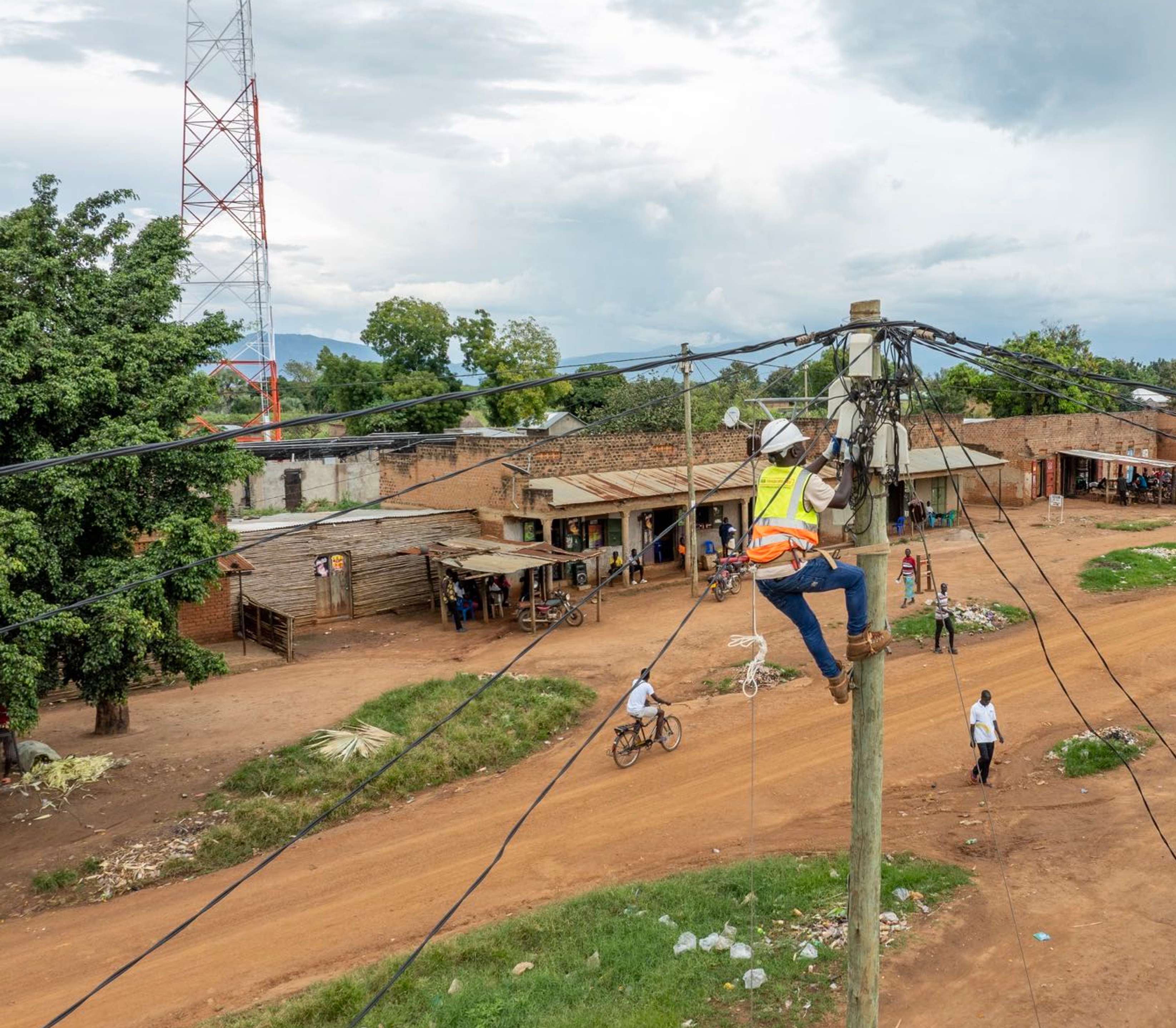A line worker in a safety vest works on a utility pole in a rural village with dirt roads, simple buildings, and a distant cell tower under a cloudy sky.
