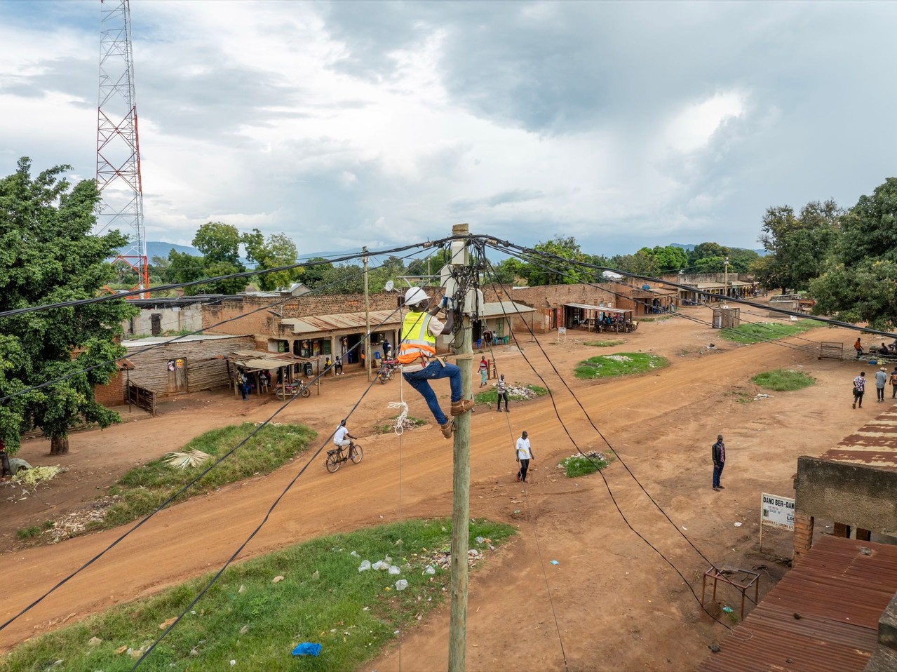 A worker in safety gear works on a utility pole in a rural village, with a communication tower in the distance.