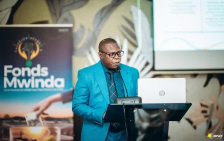 A man in a blue suit and glasses speaks at a podium with a laptop, a 'Fonds Mwinda' banner and a projection screen in the background.