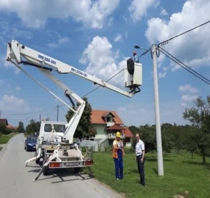 A worker in a bucket truck fixes a street light, with two colleagues standing on the grass beside the road.