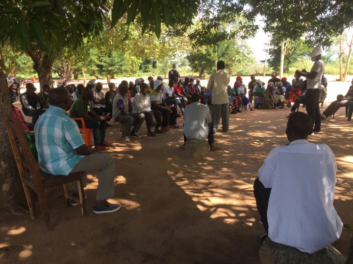 Community meeting taking place outdoors under the shade of trees.