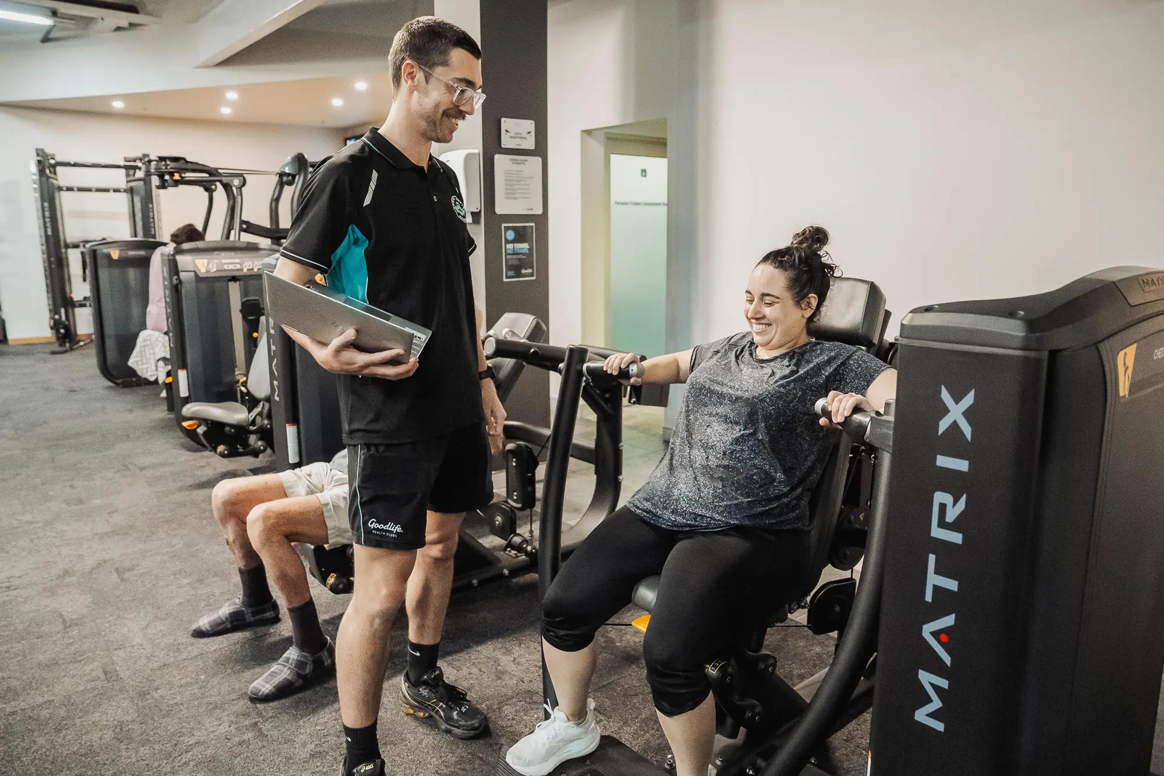 Exercise physiologist helping client with strength training, both smiling in a bright, modern gym environment.