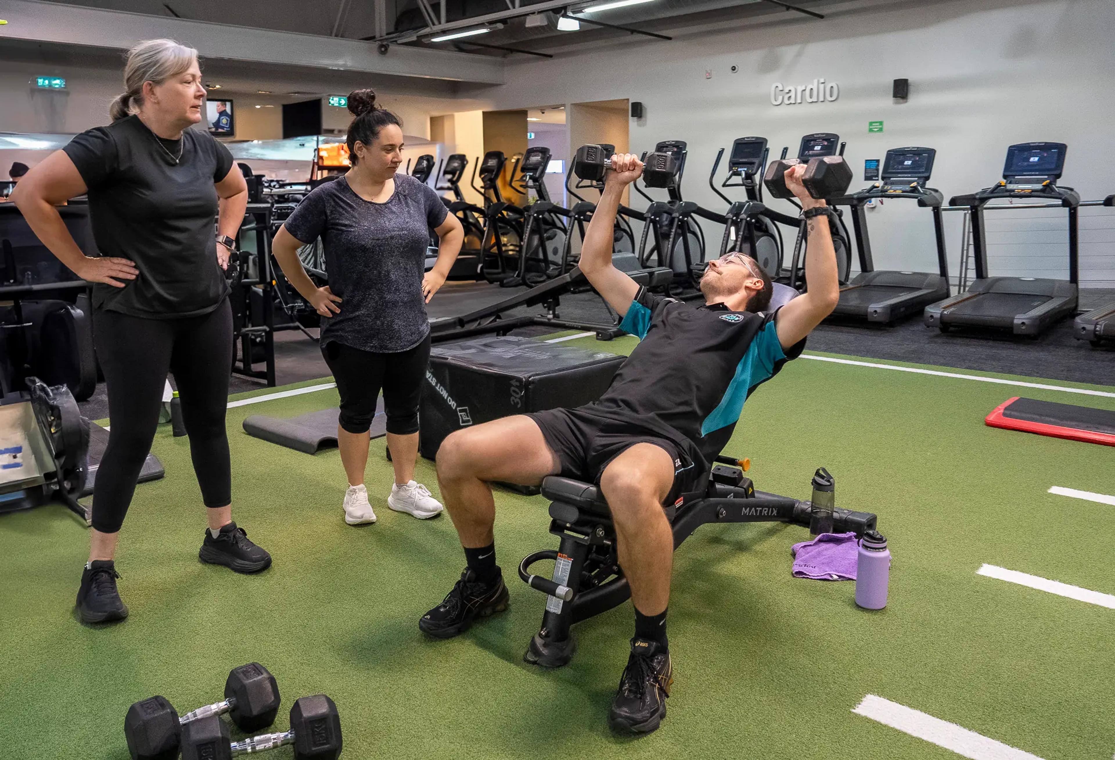 Focused trainer demonstrates proper dumbbell chest press technique while two attentive colleagues provide supportive guidance in a professional gym setting.