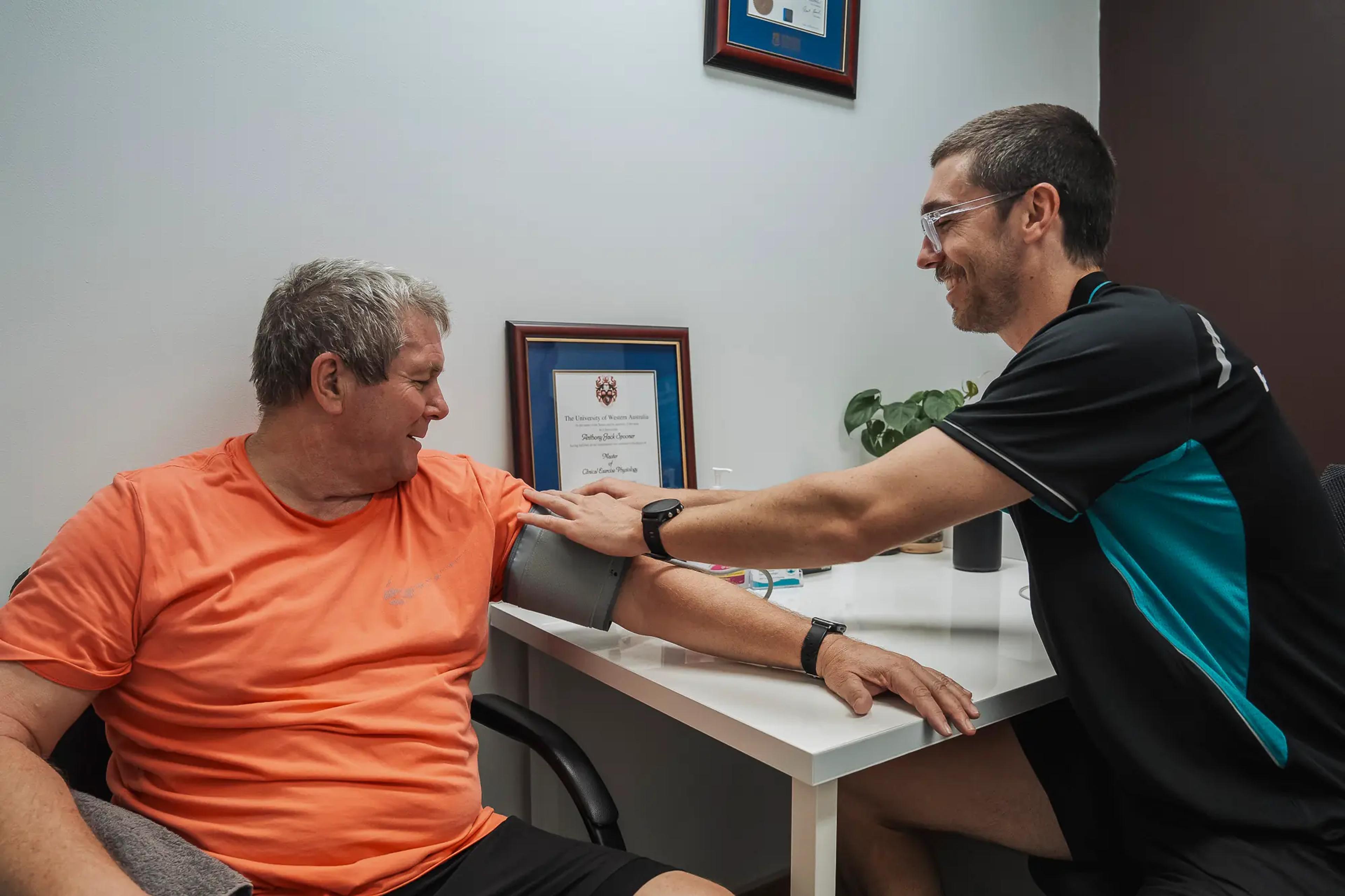 Exercise physiologist taking patient's blood pressure, both smiling warmly in clinical consultation.