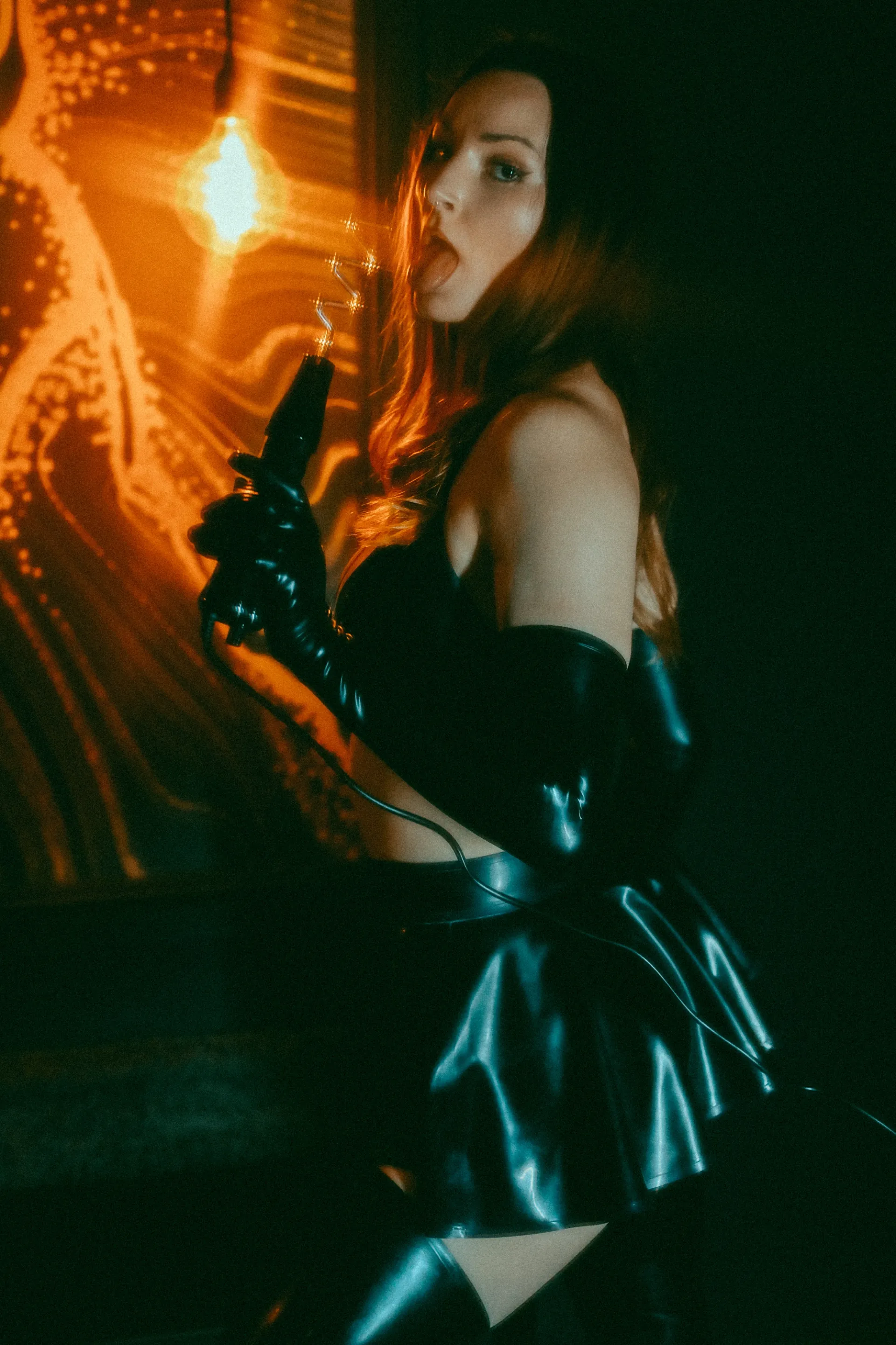 Jennifer Hex, dressed in latex, holding an electro wand in her mouth by a light.