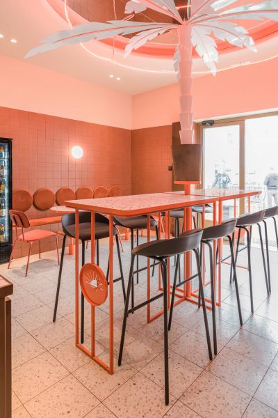 Moderný bar s vysokými čiernymi stoličkami Team pri úzkom oranžovom stole v pastelovom interiéri