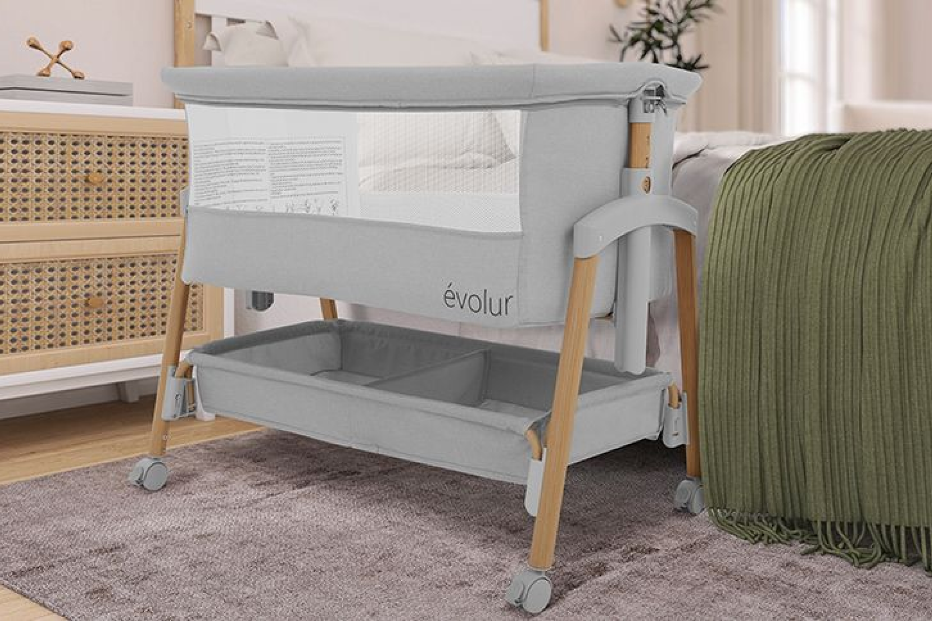 Evolur Lullaby bassinet with mesh sides and wooden legs on wheels, placed beside a bed with a green throw blanket in a cozy, modern bedroom setting.