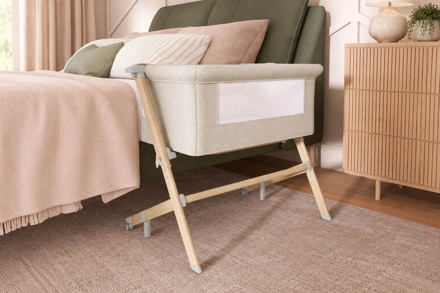 Beige bedside bassinet with wooden legs beside a blush and sage bed in a cozy, neutral-toned bedroom
