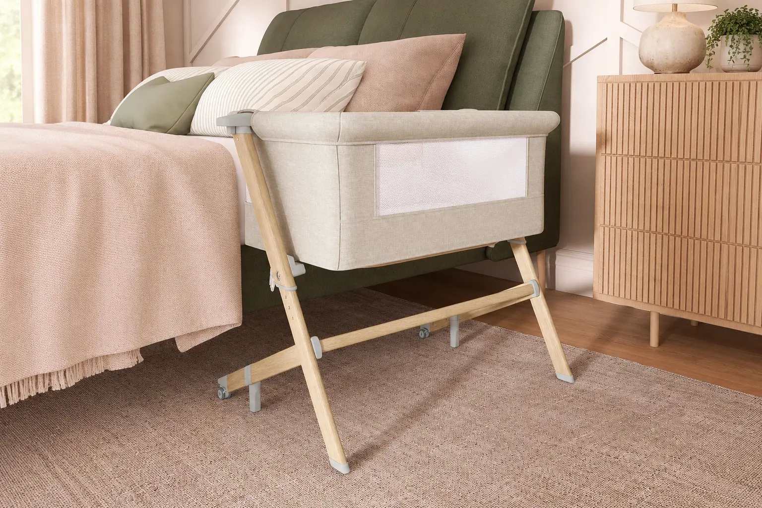 Beige bedside bassinet with wooden legs beside a blush and sage bed in a cozy, neutral-toned bedroom