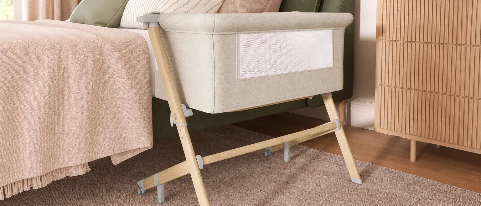 Beige bedside bassinet with wooden legs beside a blush and sage bed in a cozy, neutral-toned bedroom