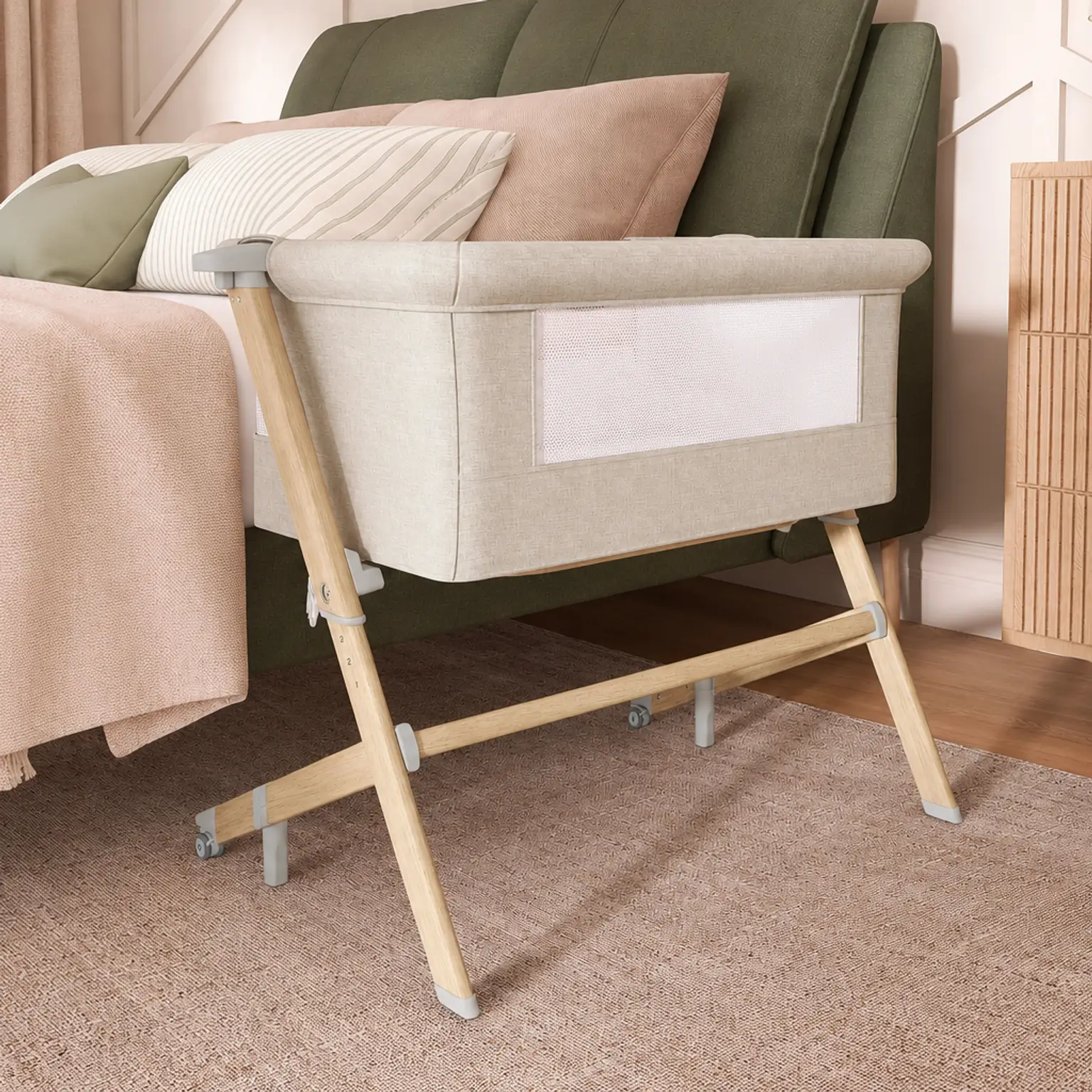 Beige bedside bassinet with wooden legs beside a blush and sage bed in a cozy, neutral-toned bedroom