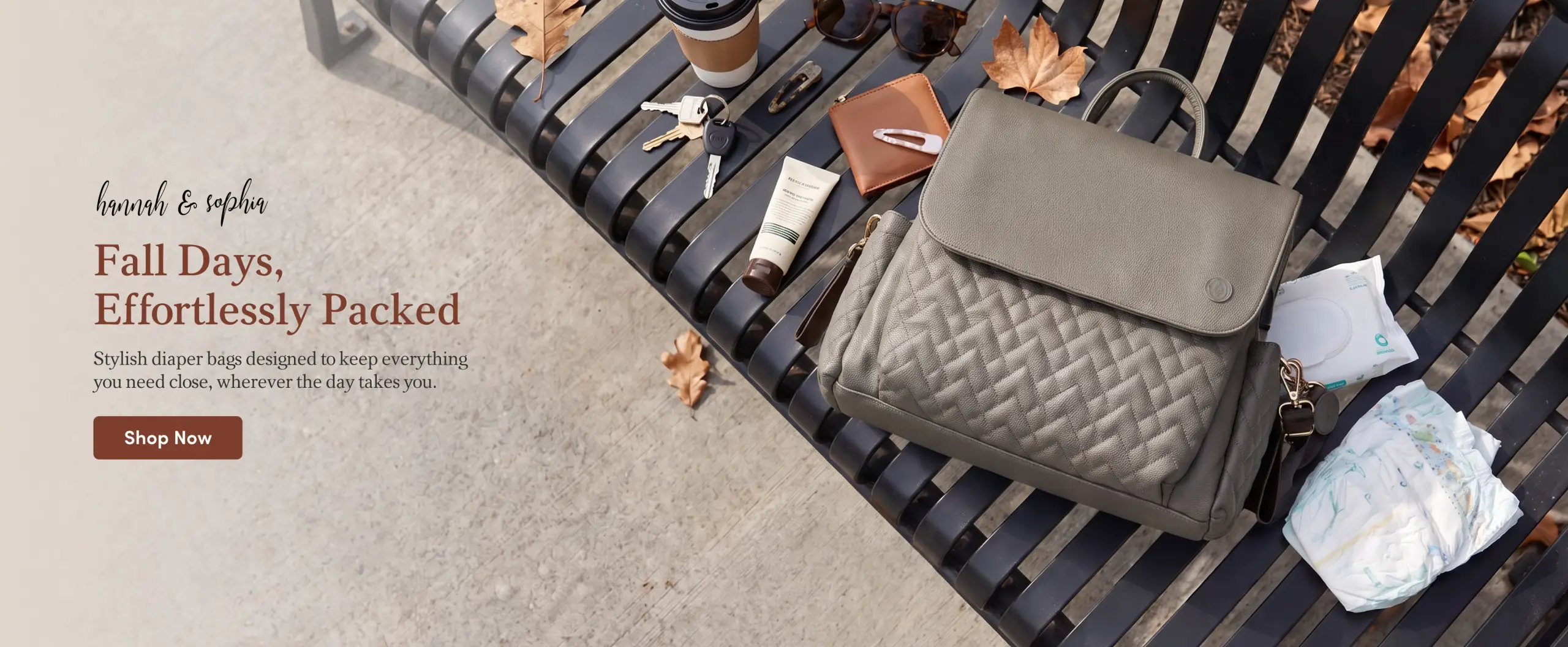 A gray quilted diaper bag sits on a black park bench with fallen autumn leaves. Nearby are a coffee cup, keys, wallet, sunglasses, lotion, baby wipes, and a diaper. Text overlay: "hannah & sophia. Fall Days, Effortlessly Packed. Stylish diaper bags designed to keep everything you need close, wherever the day takes you. Shop Now."